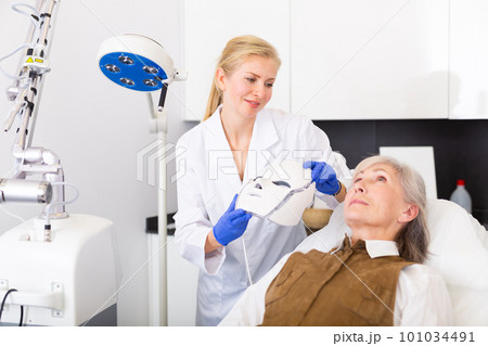 Cosmetologist using spectrum mask on elderly woman 101034491