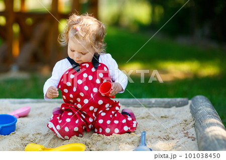 Cute toddler girl playing in sand on outdoor playground. Beautiful baby in red gum trousers having fun on sunny warm summer day. Child with colorful sand toys. Healthy active baby outdoors plays games 101038450