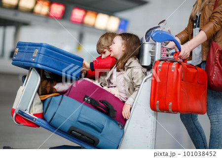 Two little kids, boy and girl, siblings and mother at the airport. Children, family traveling, going on vacation by plane and waiting on trolley with suitcases pushing by woman at terminal for flight. Two little kids, boy and girl, siblings and mother at the airport. Children, family traveling, going on vacation by plane and waiting on trolley with suitcases pushing by woman at terminal for flight. 101038452