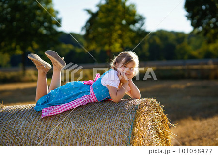 Cute little kid girl in traditional Bavarian costume in wheat field. Happy child with hay bale during Oktoberfest in Munich. Preschool girl play at hay bales during summer harvest time in Germany. Cute little kid girl in traditional Bavarian costume in wheat field. Happy child with hay bale during Oktoberfest in Munich. Preschool girl play at hay bales during summer harvest time in Germany. 101038477