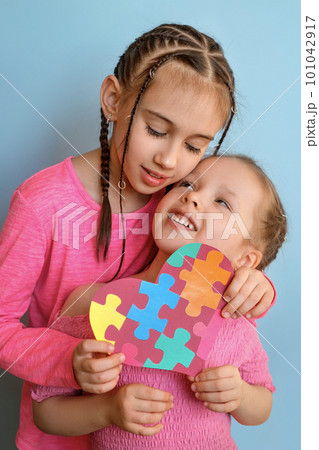 Girl gently hugs sister with autism syndrome, holding heart made of multicoloured puzzles in her hands as a sign of solidarity and unity with people suffering from autism spectrum disorder Girl gently hugs sister with autism syndrome, holding heart made of multicoloured puzzles in her hands as a sign of solidarity and unity with people suffering from autism spectrum disorder 101042917