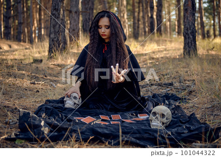 Witch in black dress with cape and hood. Posing in pine forest. Sitting on dark blanket whith fortune-telling cards and skulls on it. Full length. 101044322