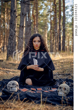 Witch in black dress with cape and hood. Posing in pine forest. Sitting on dark blanket whith fortune-telling cards and skulls on it. Full length. Witch in black dress with cape and hood. Posing in pine forest. Sitting on dark blanket whith fortune-telling cards and skulls on it. Full length. 101044325