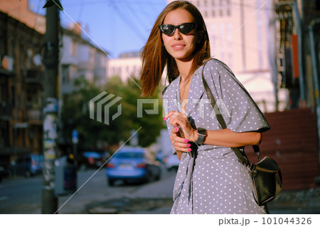 Blonde girl in long blue dress, sunglasses and a small black handbag is holding her smartphone while walking alone in the city. Close-up. Blonde girl in long blue dress, sunglasses and a small black handbag is holding her smartphone while walking alone in the city. Close-up. 101044326