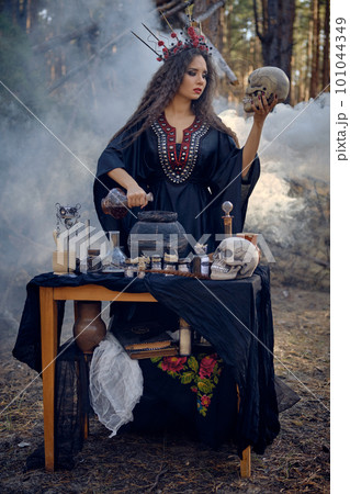 Witch in black, long dress, with red crown in hair. Posing in pine forest, holding skull, making potion. Spells and witchcraft. Full length, smoke. Witch in black, long dress, with red crown in hair. Posing in pine forest, holding skull, making potion. Spells and witchcraft. Full length, smoke. 101044349