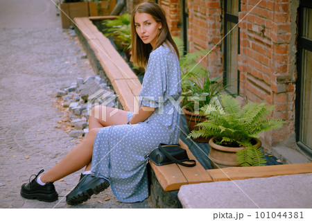 Blonde girl in long blue dress and small black handbag on her shoulder is posing sitting sideways on a parapet. Fashion and style. Full length shot. 101044381
