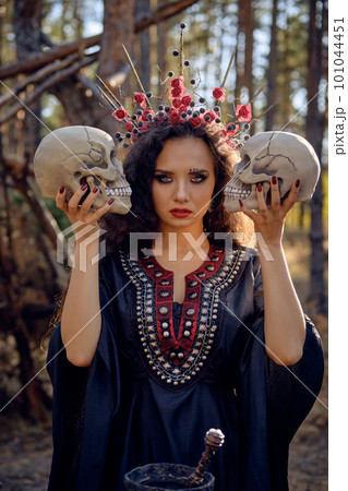 Witch in black, long dress, with red crown in her long, curly hair. Posing in pine forest. Holding skulls. Spells, magic and witchcraft. Close-up. Witch in black, long dress, with red crown in her long, curly hair. Posing in pine forest. Holding skulls. Spells, magic and witchcraft. Close-up. 101044451