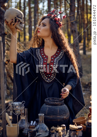 Witch in black, long dress, with red crown in her long, curly hair. Posing in pine forest. Holding skull. Spells, magic and witchcraft. Close-up. 101044472