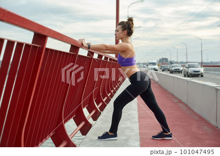Girl athlete stretching in the morning outside, in the city, on modern bridge crossing river. Active healthy lifestyle. Sport and recreation concept. 101047495