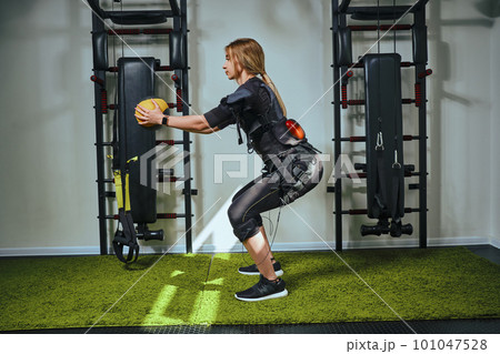 Beautiful blonde woman wearing in sensors and belts, fastens with velcro is practicing EMS fitness in a gym, building up muscles with a weighted ball. Modern kind of sport. Beautiful blonde woman wearing in sensors and belts, fastens with velcro is practicing EMS fitness in a gym, building up muscles with a weighted ball. Modern kind of sport. 101047528