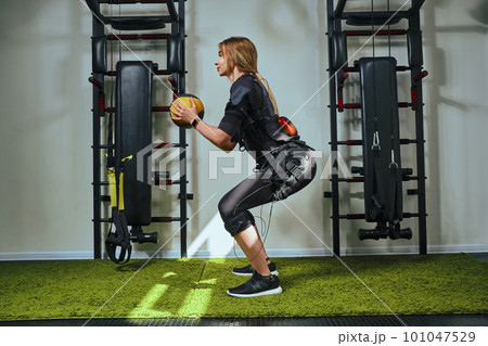 Beautiful blonde woman wearing in sensors and belts, fastens with velcro is practicing EMS fitness in a gym, building up muscles with a weighted ball. Modern kind of sport. Beautiful blonde woman wearing in sensors and belts, fastens with velcro is practicing EMS fitness in a gym, building up muscles with a weighted ball. Modern kind of sport. 101047529