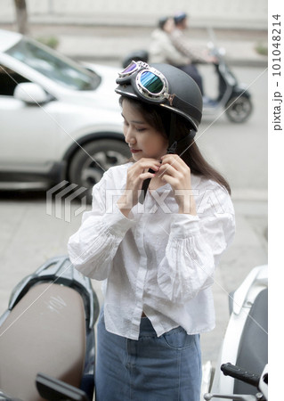 Pretty girl fastening her helmet next to a scooter on the sidewalk 101048214