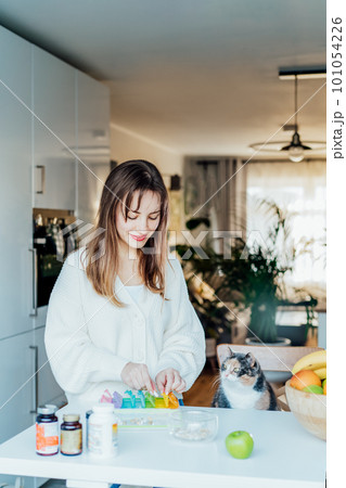 Woman sorting pills into pills organizer with her cat on the kitchen. Pill box with daily doses of medicines. Antioxidant diet vitamin supplements for health and beauty care, biohacking concept. Woman sorting pills into pills organizer with her cat on the kitchen. Pill box with daily doses of medicines. Antioxidant diet vitamin supplements for health and beauty care, biohacking concept. 101054226