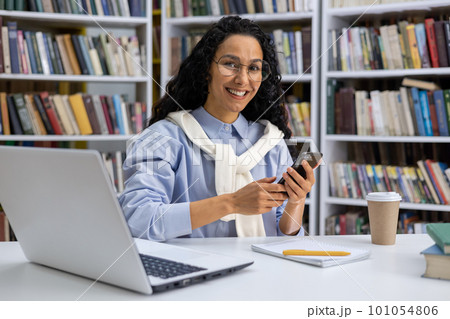Portrait of a beautiful Hispanic female student in a university library among books, a woman with curly hair is smiling and looking at camera, holding hone, using an application online learning. Portrait of a beautiful Hispanic female student in a university library among books, a woman with curly hair is smiling and looking at camera, holding hone, using an application online learning. 101054806