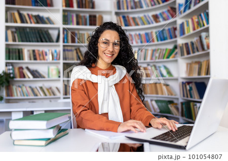 Portrait of cheerful smiling girl student, woman looking at camera sitting studying with laptop, Hispanic woman wearing glasses studying online in university library among glasses using laptop. 101054807