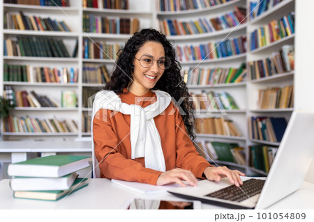 Beautiful Latin American student studying in university library, woman with curly hair and glasses typing on laptop keyboard smiling, having fun preparing for exam. Beautiful Latin American student studying in university library, woman with curly hair and glasses typing on laptop keyboard smiling, having fun preparing for exam. 101054809