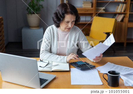 Middle aged senior woman sit with laptop and paper document. Pensive older mature lady reading paper bill pay online at home managing bank finances calculating taxes planning loan debt pension payment 101055001
