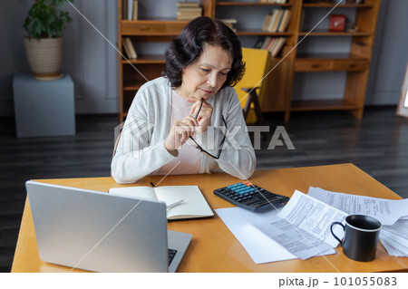 Middle aged senior woman sit with laptop and paper document. Pensive older mature lady reading paper bill pay online at home managing bank finances calculating taxes planning loan debt pension payment 101055083
