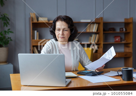 Middle aged senior woman sit with laptop and paper document. Pensive older mature lady reading paper bill pay online at home managing bank finances calculating taxes planning loan debt pension payment 101055084