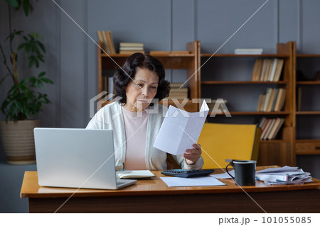 Middle aged senior woman sit with laptop and paper document. Pensive older mature lady reading paper bill pay online at home managing bank finances calculating taxes planning loan debt pension payment 101055085