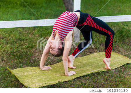 Full length of young adult attractive yogi woman practicing yoga, stretching in wheel exercise, urdhva dhanurasana pose, working out on mat outdoor in green park, wearing sportswear. Full length of young adult attractive yogi woman practicing yoga, stretching in wheel exercise, urdhva dhanurasana pose, working out on mat outdoor in green park, wearing sportswear. 101056130