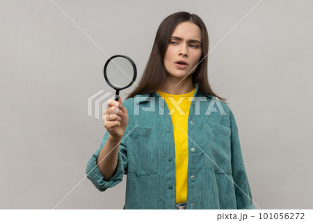 Curious woman with dark hair looking away through magnifying glass, spying, finding out something, inspecting, wearing casual style jacket. Indoor studio shot isolated on gray background. 101056272