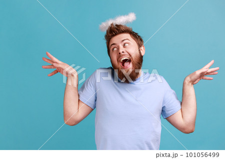 Portrait of handsome funny bearded angelic man with holy nimbus on head standing with raised hands and looking away with open mouth. Indoor studio shot isolated on blue background. Portrait of handsome funny bearded angelic man with holy nimbus on head standing with raised hands and looking away with open mouth. Indoor studio shot isolated on blue background. 101056499
