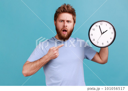 Portrait of shocked astonished surprised handsome bearded man pointing at wall clock, deadline, having puzzled facial expression. Indoor studio shot isolated on blue background. 101056517