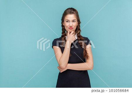Portrait of cunning woman with braids standing holding chin pondering, thinking about devil plan or prank, wearing black dress. woman Indoor studio shot isolated on blue background. 101056684