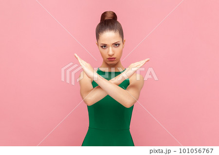 Portrait of attractive strict serious woman with bun hairstyle standing and showing x sign, no way gesture, prohibition, wearing green dress. Indoor studio shot isolated on pink background. 101056767