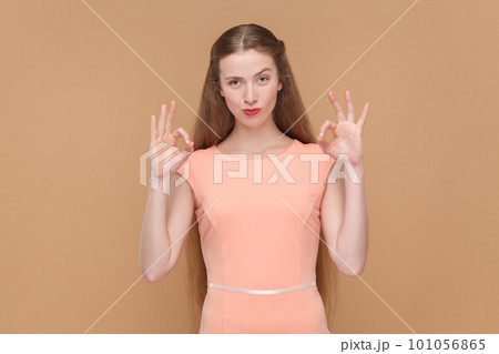 Portrait of adorable positive woman with long hair standing showing okay sign, approval gesture, like service, wearing elegant dress. Indoor studio shot isolated on brown background. 101056865