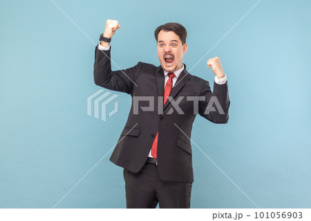 Portrait of excited extremely happy man standing with clenched fists, celebrating his victory, screaming hurray, wearing black suit with red tie. Indoor studio shot isolated on light blue background. Portrait of excited extremely happy man standing with clenched fists, celebrating his victory, screaming hurray, wearing black suit with red tie. Indoor studio shot isolated on light blue background. 101056903
