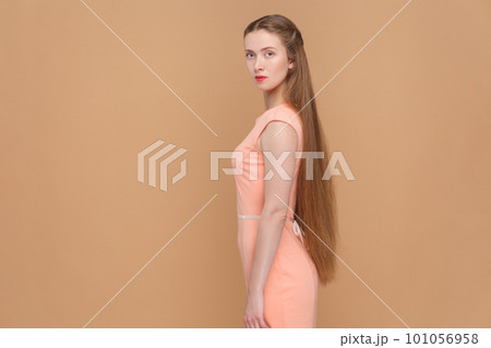 Side view portrait of serious strict woman with long hair looking at camera with bossy expression, being in bad mood, wearing elegant dress. Indoor studio shot isolated on brown background. 101056958