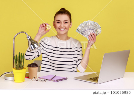 Satisfied woman office worker holding and showing fan of hundred dollar banknotes and bitcoin crypto coin looking at camera with toothy smile. Indoor studio studio shot isolated on yellow background. 101056980