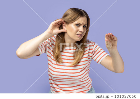 Portrait of blond woman wearing striped T-shirt holding hand near ear trying to listen quiet conversation, overhearing gossip, showing blah gesture. Indoor studio shot isolated on purple background. Portrait of blond woman wearing striped T-shirt holding hand near ear trying to listen quiet conversation, overhearing gossip, showing blah gesture. Indoor studio shot isolated on purple background. 101057050