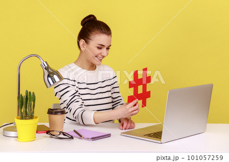 Satisfied woman sitting at workplace, showing big red hashtag symbol to laptop screen, sharing viral content, tagged message. Indoor studio studio shot isolated on yellow background. Satisfied woman sitting at workplace, showing big red hashtag symbol to laptop screen, sharing viral content, tagged message. Indoor studio studio shot isolated on yellow background. 101057259