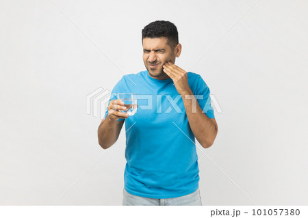 Portrait of unhealthy ill unshaven man wearing blue T- shirt standing with glass of cold water, feels pain in his tooth, has sensitive teeth. Indoor studio shot isolated on gray background. Portrait of unhealthy ill unshaven man wearing blue T- shirt standing with glass of cold water, feels pain in his tooth, has sensitive teeth. Indoor studio shot isolated on gray background. 101057380