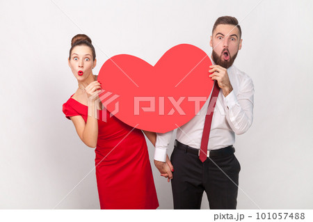 Portrait of amazed shocked man in white shirt and woman in red dress standing together, peeping out from big heart, expressing love. Indoor studio shot isolated on gray background. Portrait of amazed shocked man in white shirt and woman in red dress standing together, peeping out from big heart, expressing love. Indoor studio shot isolated on gray background. 101057488