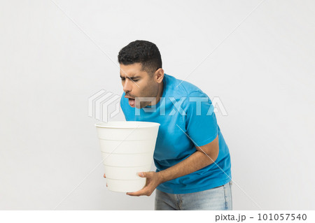 Portrait of sick unhealthy ill unshaven man wearing blue T- shirt standing suffering stomachache, feels nausea and vomits, holding bin in hands. Indoor studio shot isolated on gray background. 101057540