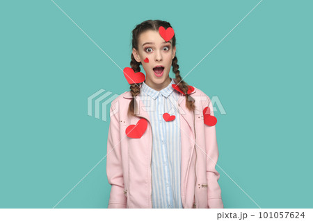 Portrait of amazed surprised teenager girl with braids wearing pink jacket standing covered with little red hears, looking at camera with open mouth. Indoor studio shot isolated on green background. Portrait of amazed surprised teenager girl with braids wearing pink jacket standing covered with little red hears, looking at camera with open mouth. Indoor studio shot isolated on green background. 101057624