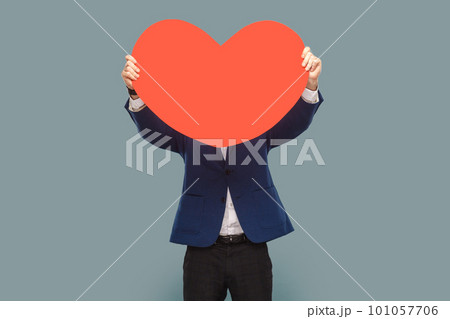Unknown man standing hiding his face behind big red heart, being shy to say about his feelings, wearing official style suit. Indoor studio shot isolated on light blue background. 101057706