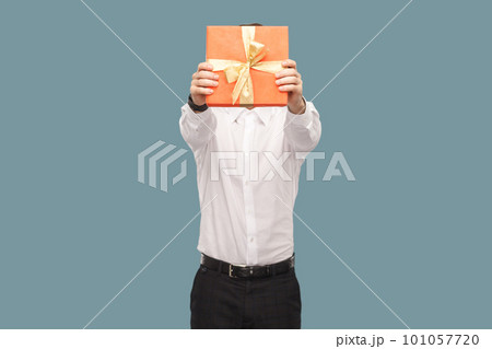 Portrait of unknown anonymous man holding out and covering his face with red gift box, giving present for anniversary, wearing Indoor studio shot isolated on light blue background. 101057720