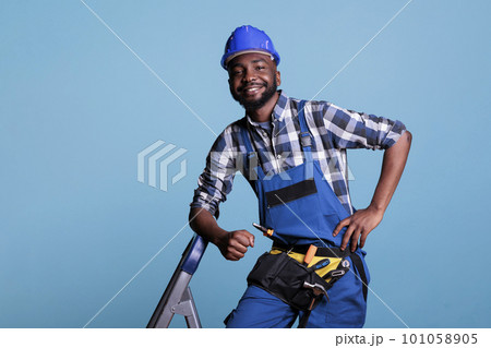 Optimistic builder looking at camera with big smile while resting after finishing work in studio shot against blue background. Contractor using tool belt needed for renovation construction tasks. 101058905