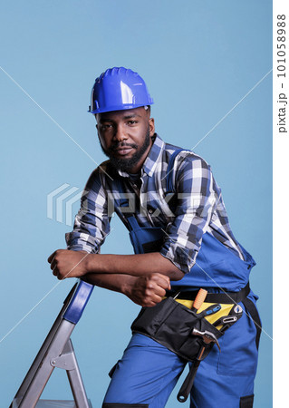 Builder looking calm leaning against ladder in studio shot against blue background. African american construction worker wearing hard hat and work uniform looking into camera. 101058988