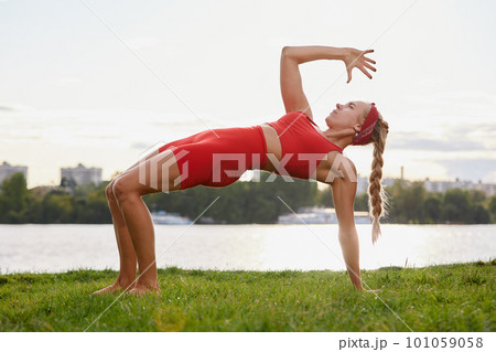 A woman is engaged in animal flow gymnastics, close-up outdoors near the river on green grass in red clothes. A woman is engaged in animal flow gymnastics, close-up outdoors near the river on green grass in red clothes. 101059058
