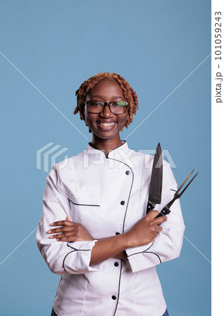 African american female cooker, curly haired posing with professional kitchen knife, sharpener. Smiling afro chef holding kitchen utensils. Portrait of uniformed young female restaurant worker. 101059243