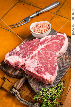 Porterhouse raw beef meat Steak, T-bone with thyme and sea salt on a wooden board. Orange background. Top view 101070892