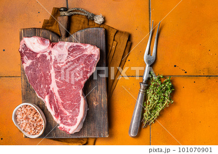 Porterhouse raw beef meat Steak, T-bone with thyme and sea salt on a wooden board. Orange background. Top view 101070901