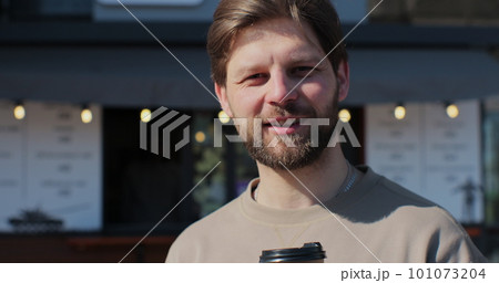 Handsome bearded man holding cup of take-out coffee and smiling standing on street on nice day, drinking coffee. 101073204