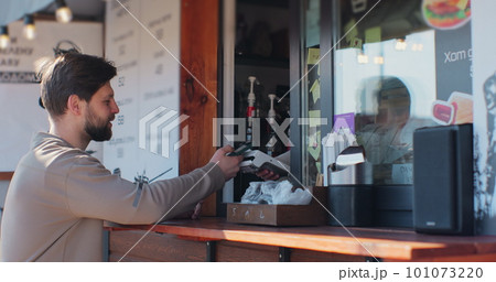 Young man is buying takeaway coffe in coffee-shop and paying with smartphone making contactless payment. Modern technology and banking concept. Young man is buying takeaway coffe in coffee-shop and paying with smartphone making contactless payment. Modern technology and banking concept. 101073220
